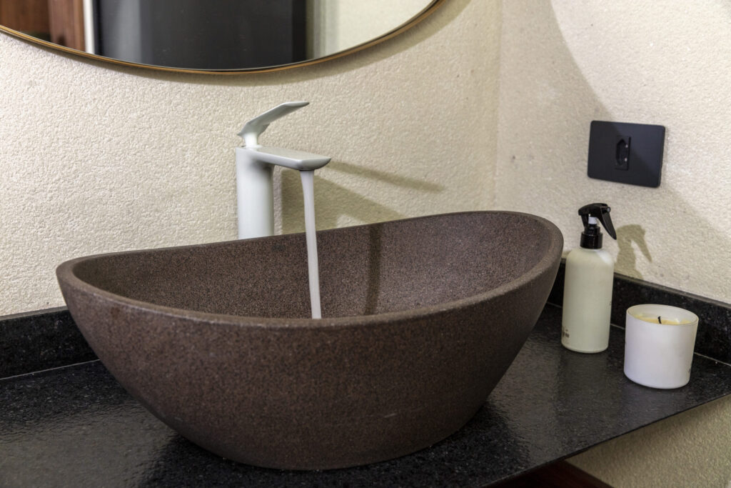 cuba duplo arco geostone dark red terrazzo em cima de uma bancada preta. o monocomando que compõem o lavabo é rainbow matt white. ao lado da cuba tem uma vela aromática e um spray, ambos na cor branca. este lavabo possui as dimensões ideais para um banheiro.